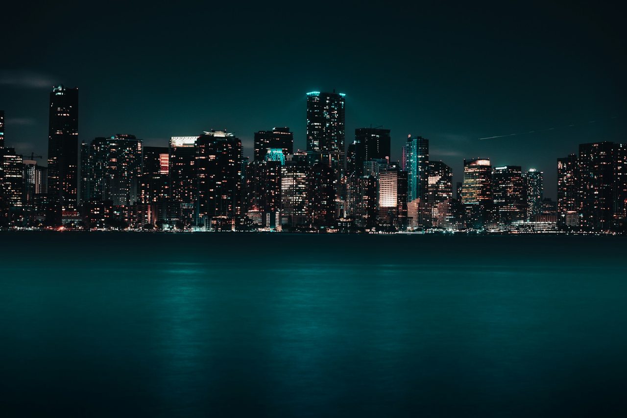 Temporary design photograph of a city skyline at night, with illuminated high-rise buildings across a dark shoreline and at least one construction crane. Not a photograph of an ACI Capital Partners property.